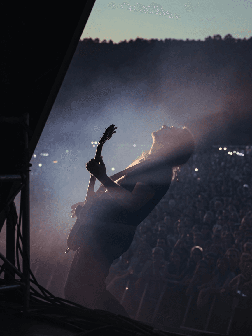 A guitarist backlit through stage haze from the side wing at dusk