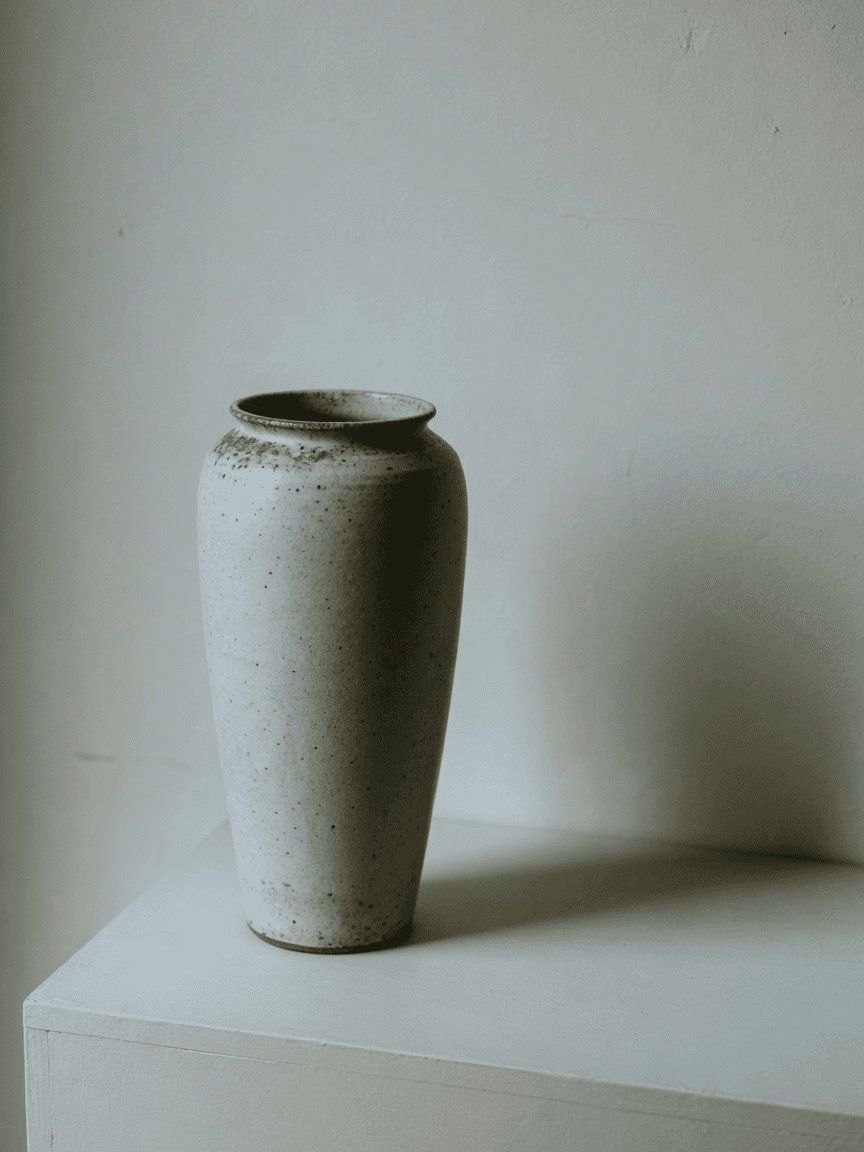 A tall ash-glazed stoneware vessel on a plinth against a bare wall